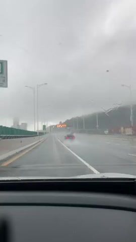 The video shows a view from inside a car driving on a wet highway during what appears to be a rainstorm. The windshield has raindrops on it, and the road surface is visibly wet, with water splashing from a car ahead. A red car is in the distance, moving away from the camera. There are streetlights and signs visible along the highway, and on the left side, there's a barrier with green posts and what looks like buildings or structures beyond it. The sky is overcast and gray.