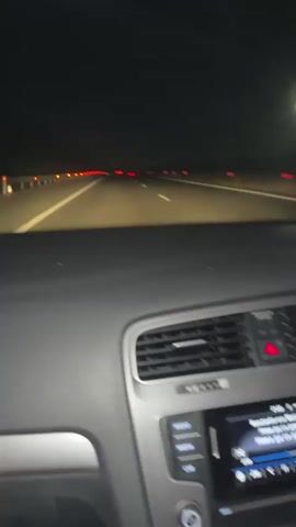 The video shows the dashboard of a car moving on a highway at night. The highway is dark but the car headlights and taillights of other vehicles are visible. Inside the car, the dashboard, air vents, and the infotainment system can be seen. The driver's hands are visible moving the steering wheel. The steering wheel has a shiny, possibly rhinestone-studded, cover. A silver bracelet is visible on the driver's wrist. An air freshener with a "VIP" design hangs from the rearview mirror. Music is playing.