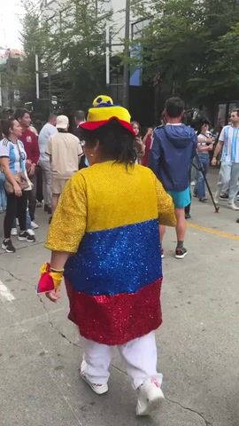 A group of people are gathered on a street. In the foreground, a person wearing a sequined jacket in the colors of the Colombian flag, a yellow hat with blue and red accents, and white pants walks away from the camera. To the right of this person, a woman wearing a yellow shirt with blue pants and a brown bag over her shoulder walks in the same direction. Other individuals, some wearing similar team colors, are also present, and one person is holding a small flag. The environment appears to be an urban street with buildings and trees visible in the background. There are also large sculptures present, including a statue of a person in athletic attire and a large soccer ball. The atmosphere seems festive.