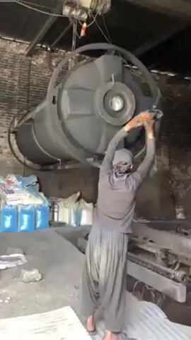 A man is pulling on a rope that is attached to a large, circular, industrial object suspended from the ceiling by a motor. The object is mostly black with a gray center and is hanging in front of a brick wall. The man is wearing a gray long-sleeve shirt, baggy black pants, a hat, and a face covering. He is barefoot. To the left of the man are several blue and white bags. To the right is a metal structure with rollers. The floor is cluttered with debris, including a piece of corrugated metal. The scene appears to be in an industrial setting.