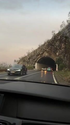 The video shows a view from inside a car driving into a tunnel. The road is wet, indicating rain or recent rain. Several cars are visible on the road ahead, driving into the tunnel. The tunnel is illuminated by lights. The surrounding environment appears to be mountainous, with rocky slopes visible on either side of the tunnel entrance. The sky is overcast.