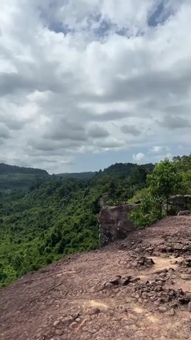 The video pans across a lush, green landscape filled with rolling hills and dense vegetation under a cloudy sky. The foreground is dominated by a rocky, reddish-brown cliff face. In the distance, more hills covered in trees stretch out towards the horizon. The scene is devoid of people and any discernible action, offering a static view of a natural environment.