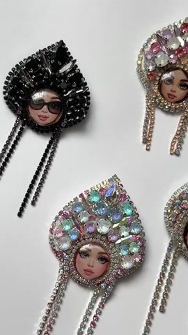 The video shows a top-down view of four brooches arranged on a white surface. Each brooch features a small, illustrated face of a woman, adorned with a large, ornate headdress made of rhinestones and colorful jewels. Dangling from the headdresses are multiple strands of smaller rhinestones. The brooches are positioned in a rough diamond formation, with one on the top left, two in the middle row, and one partially visible on the bottom right. The brooches vary in color and jewel arrangement, with one on the left being predominantly black and featuring sunglasses on the illustrated face. The lighting is bright, causing reflections on the jewels.