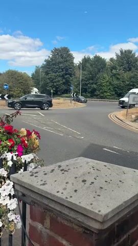 The video shows a street scene with a roundabout. In the foreground, there is a brick pillar with a stone cap covered in dark spots, and a planter filled with colorful flowers, including red, pink, and white blooms. Beyond the pillar, a road curves into a roundabout. Several vehicles are visible on or approaching the roundabout. A white van is traveling clockwise on the roundabout, and a dark-colored car is also on the roundabout, moving in the same direction. Further down the road, another white van is parked on the side. On the left side of the roundabout, a dark grey car is waiting at an intersection. The sky is blue with scattered white clouds, and green trees line the background. The lighting suggests it is daytime.