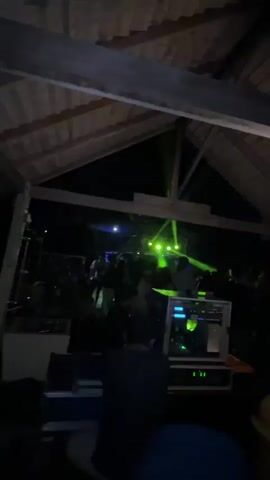 The video shows an indoor party scene at night, likely a bar or club. Green and blue lights flash from the stage at the back, illuminating a crowd of people dancing. In the foreground, a person is seen operating DJ equipment. A table in the lower right corner holds several bottles of alcohol and a few other items. The ceiling is made of exposed wooden beams. The overall atmosphere appears to be energetic and festive.