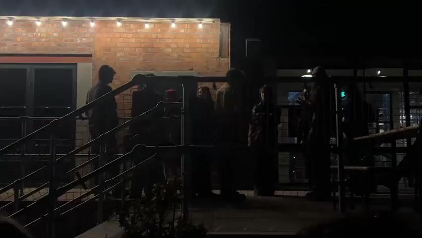 The video captures an evening scene on an outdoor patio or rooftop in Dundee, United Kingdom. Approximately ten to twelve people are gathered, mostly visible as silhouettes against the ambient light. They appear to be socializing and conversing in small groups.

The setting includes a red brick building on the left, with string lights strung along its upper edge, providing warm illumination to the immediate area. A dark metal railing system is prominent in the foreground and midground, suggesting a balcony or stairway. The background is dark, indicating night, with faint artificial lights visible in the distance, hinting at an urban environment. A small plant can be seen at the bottom of the frame. The overall atmosphere seems relaxed and social. No visible text is present in the video.