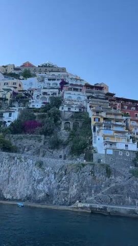 The video shows a view of a coastal town built on a steep hillside, with buildings cascading down towards the water. The architecture is characterized by colorful buildings with balconies and terraces, interspersed with lush greenery and rocky cliffs. The water in the foreground is a deep blue, with several boats of various sizes docked or moving in the harbor. People are visible walking along the waterfront, some seated at outdoor cafes. The sky is clear and blue, indicating fair weather. The scene appears to be a popular tourist destination.