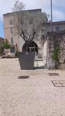 This video pans across a sunlit town square in Cahors, France. The scene is bustling with activity, suggesting a lively midday or early afternoon. The weather appears to be pleasant, with a clear blue sky dotted with a few wispy clouds, casting bright shadows.

The architecture is a blend of old and new. We see historic stone buildings with arched doorways and balconies, interspersed with more modern structures. The ground is paved with cobblestones, typical of European towns. In the foreground, there are wooden benches, some occupied by people sitting or walking, and a large planter with a green shrub. Further back, a cafe with outdoor seating is visible, with patrons enjoying the atmosphere.

A prominent architectural feature is a grand stone building with a large arched entrance. Above the arch, the text "Révolution R" is displayed, indicating a business or establishment. Further along, another building features a storefront with large windows. Reflected in one window is the text "CLOS CHAI RESTAURANT BAR A VIN CAFE", identifying it as a restaurant and wine bar.

The presence of parked motorcycles and cars suggests everyday life and local commerce. The overall mood is relaxed and pleasant, capturing the essence of a European town square on a beautiful day. The video shows people engaged in casual activities, enjoying the outdoor environment and the amenities offered by the local businesses.