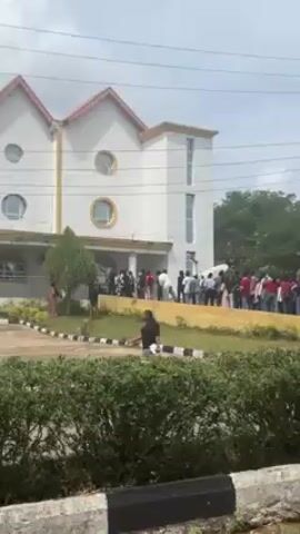 The video depicts a crowd of people outside a building, likely a university or college, as suggested by the architecture of the building and the age range of the individuals present. There's a long line of people queued up in front of the building at the start. The camera then pans around the location, showing groups of people standing and walking around on the roads and walkways. There's greenery and trees scattered throughout the area. The sky is partially cloudy. People are dressed in casual clothing.