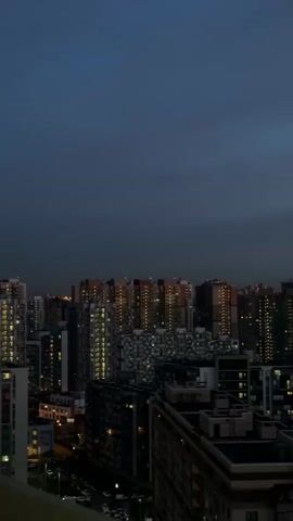 The video shows an aerial view of a city skyline at dusk. Numerous high-rise buildings dominate the view, with many windows lit up, creating a grid-like pattern. The sky is a dark blue, indicating the early evening hour. A few cars can be seen on the streets below. No people are visible in the frame.