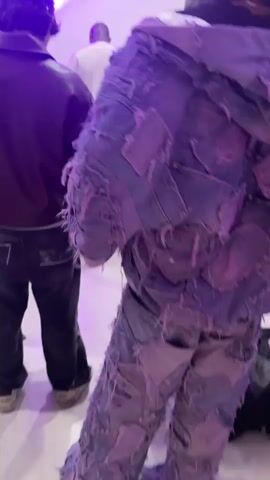 The video shows the back view of several people in what seems to be a photo studio. The first person is wearing a jacket and pants, both made of patched and distressed blue denim. The second person wears a black hooded jacket made of a similar patchwork pattern. The scene shifts to show another person in a brown leather jacket and a black studded cap. The environment includes photography equipment like lights and reflectors, a ladder, and a neon sign. Some people are moving around, possibly setting up or working on a photoshoot. The overall lighting is purple-toned.