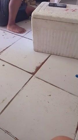A top-down shot reveals a person sitting near a white cooler on a tiled floor. The person is barefoot and wearing dark shorts or pants. The cooler has some writing on it in red. A smartphone is lying on top of the cooler. The tiles are off-white and appear somewhat dirty. The camera is held by a person, and their arm is visible in the upper right of the frame.