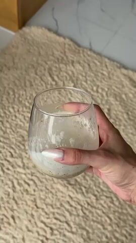 A hand with manicured nails holds a clear glass filled with a frothy, light-colored beverage. The glass has a rounded shape and is partially filled. The background features a textured, light-brown rug and a blurred tiled surface. The hand is positioned in the lower right quadrant of the frame.