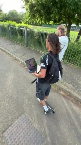 A man rides a scooter on a paved path, holding a laptop open. A woman walks beside him. The laptop screen shows a soccer game. The man is wearing a black jersey with green, white, and red details, gray shorts, and a backpack. He’s also wearing black and white shoes. The woman walking alongside him is wearing a white sweatshirt. The path is bordered by a metal fence and green foliage.
