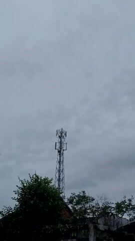 A video shows a cell tower against a cloudy sky. The tower is a lattice structure with multiple antennae mounted on it. The camera zooms in on the top of the tower, then zooms back out. The tower is framed by trees and a fence.