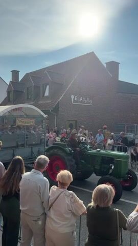 A tractor pulls a trailer filled with people on a street lined with buildings. Several people stand on the sidewalk watching the parade. The sun is shining brightly.