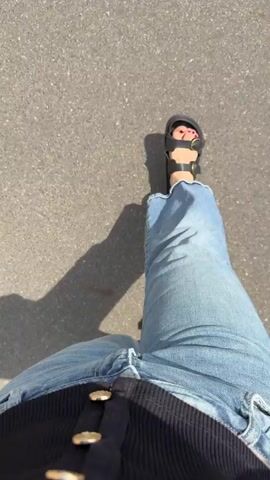 The video displays a first-person perspective of someone walking on a paved surface. The individual is wearing wide-leg denim jeans and black sandals with gold buckles. Their toenails are painted red. Sunlight casts a shadow of the person on the ground. The environment appears to be outdoors, possibly a street or sidewalk, with the texture of asphalt visible. The action is solely the person's gait.