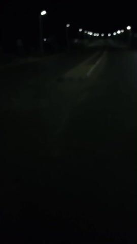 A shaky, low-resolution video shows a dark road at night, illuminated by spaced-out streetlights. The camera seems to be moving forward. There's a low divider in the middle of the road for a while. Occasionally, blurred buildings are visible in the background.
