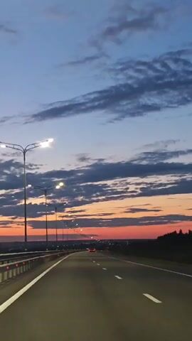 The video shows a view from a car traveling on a highway at dusk. Streetlights illuminate the road, and the sky displays a gradient of blue and orange hues as the sun sets. The road has white lane markings, and a guardrail runs along the left side. In the distance, a few cars can be seen with their headlights on. The overall scene depicts a serene and quiet journey on a highway as night approaches.