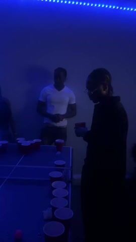 The video shows a group of people in a room lit with blue LED lights. A TV is playing a video game, possibly Mario Kart. Several people are in the foreground, with one person in a white t-shirt visible near a table with red cups on it. There's some movement and noise in the background, suggesting a casual gathering.