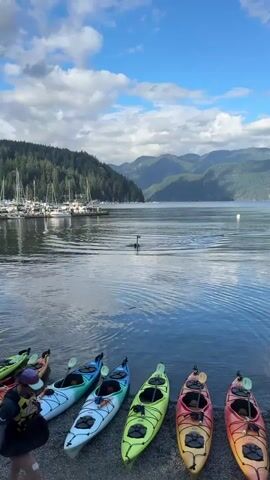 The video begins with a wide shot of a body of water, a marina with sailboats docked on the left, and a forested mountainside in the background under a cloudy blue sky. In the foreground, several colorful kayaks are lined up on the shore. A person is standing next to the kayaks, holding a black bag. The camera then zooms in on the water where a person is floating on a hydrofoil board, holding onto the board and the foil. The person is partially submerged in the water, with only their head and upper body visible. The water is calm with ripples. The scene appears to be a recreational activity on a lake or bay.