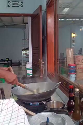 The video shows a person cooking in a kitchen. The person is using a wok and a spatula to stir food that is steaming. The wok is on a gas stove. There are bottles of sauce and other items on a shelf above the stove. The kitchen appears to be indoors.