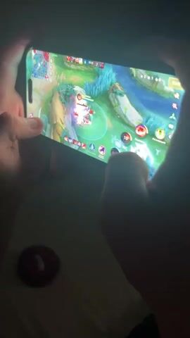 A person's hands are holding a smartphone horizontally, playing a video game. The screen displays a colorful, cartoonish landscape with a river and trees. A character in the game is surrounded by a glowing blue circle, suggesting it's a target or under some effect. The person's thumbs are on the screen, likely controlling the game. The lighting is dim, with the phone screen being the primary light source. A small, circular red object with a white center is visible on the surface below the phone.