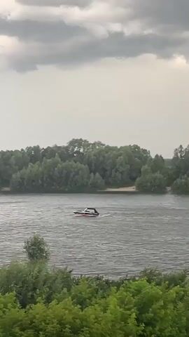 The video shows a wide shot of a river under a cloudy sky. A small, red and white motorboat is moving from left to right across the water. The riverbanks are lined with lush green trees. In the foreground, there is a dense patch of green foliage. The overall impression is of a natural landscape with a single recreational activity.