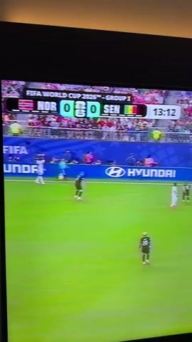 The video shows a television screen displaying a soccer match. The score is tied at 0-0 between NOR and SEN. The time on the clock is 13:16. The game is part of the FIFA World Cup 2026, Group 1. The match is being played atford, New Jersey. Players in white and black jerseys are visible on the green field. There are spectators in the background wearing pink. A blue banner with the Hyundai logo is displayed along the sideline.