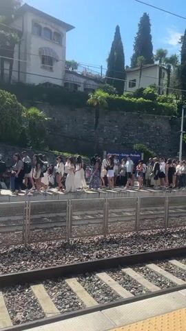 A video shows a long line of people standing on a train platform. The people are dressed in casual summer clothing and many are carrying luggage. They are all facing towards the right of the frame, as if waiting for a train. The platform has a metal fence separating it from the train tracks. In the background, there are buildings on a hillside covered in trees. The sky is blue and the sun is shining, indicating a clear day. The overall impression is one of a busy travel hub on a pleasant day.