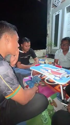 In a low-angle shot, four individuals are gathered around a small, blue, foldable table adorned with a cartoon image of Doraemon. They are seated on the floor, which is covered by a colorful, patterned mat. The scene appears to be taking place outdoors at night, with the dark sky visible in the background. A brick wall and a modern building with white doors and windows frame the setting.

The individuals are engaged in a game, possibly dominoes, as small white tiles are arranged on the table. One person in the foreground, on the left, is smoking a cigarette and actively participating in the game. Another man, wearing a black t-shirt, sits opposite him, also appearing focused on the game. A third man, wearing glasses and a grey and white striped polo shirt, is seated on the right, observing the game with a neutral expression. The fourth individual is partially visible on the far right.

The atmosphere seems casual and relaxed, suggesting a social gathering for recreation. The lighting is dim, likely from artificial sources not fully captured in the frame, casting soft shadows. The presence of cigarettes and the late hour indicate a nighttime activity. The building's architecture suggests a residential area in Indonesia, and the "Meunasah Raya" context places it within that specific location. There is no visible text in the media file that provides further context or information.
