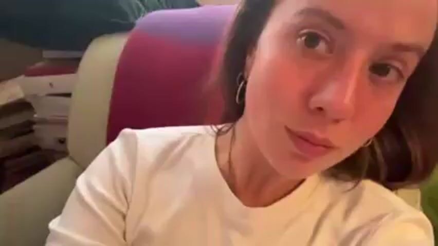 The video shows a woman with light skin and light brown hair wearing a white long-sleeved shirt. She is sitting in a light-colored chair with a striped purple and pink cushion behind her head. A stack of books is visible in the background to her left. She makes a kissing face at the camera, then smiles and winks.