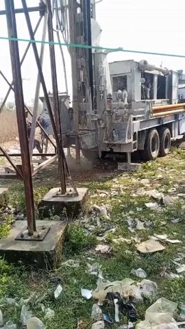 The video shows a drilling rig at work in a grassy area littered with trash. The rig is mounted on the back of a truck and is drilling a hole into the ground, with dust and debris being kicked up as it operates. To the left of the rig is a metal structure made of beams and pillars. There's a person visible sitting on the structure, observing the drilling operation. The surrounding environment has patches of green grass mixed with scattered trash. The sky is bright, suggesting it's daytime.