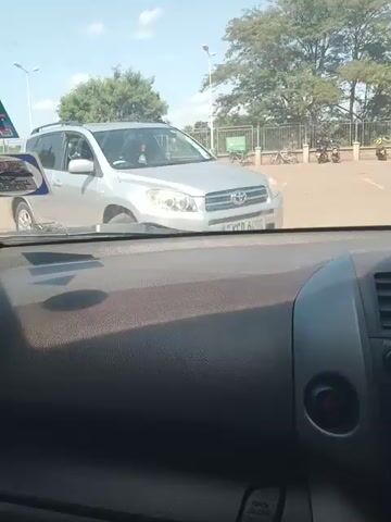 The video, taken from inside a car, shows a silver Toyota RAV4 ahead in traffic. The RAV4 has a license plate visible. A person walks behind the RAV4. The car taking the video has a black dashboard and AC vents visible. Trees and a light post are visible in the background. The sky is blue and the weather is sunny.