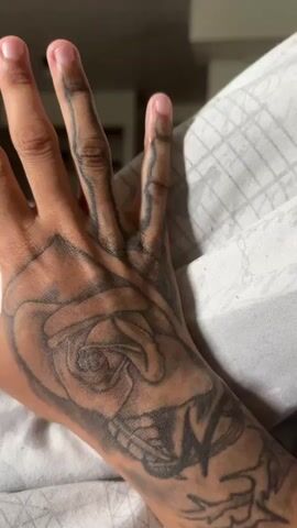 A close-up shot shows a person's hand lying on a surface, likely a bed, covered with white textured bedding. The hand has a large tattoo of a rose and leaves on the back and wrist, with thinner line tattoos extending up the fingers. The person's skin tone is light brown. The focus is on the hand and the tattoos. The background is slightly out of focus, suggesting the camera is focused on the details of the tattoo. The lighting is soft and even.