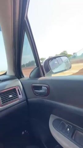 From the interior of a car with a cracked windshield, the view shows other cars driving on a dirt road under a clear blue sky with scattered clouds. The car's interior features dark gray dashboard with wood trim and a gray door panel. Other vehicles are visible on the road ahead.