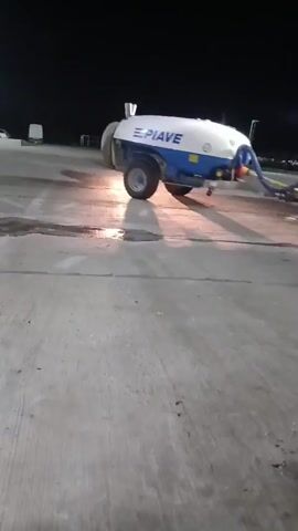 A red tractor with bright headlights pulls a white and blue tank trailer with "EPIAVE" written on it across a concrete surface at night.