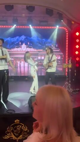 The video opens with a shaky, blurry shot of a stage lit with bright red lights. A person in a white shirt and dark pants, holding a microphone, stands on the stage. The camera then pans to the right, revealing a crowded, opulent interior with ornate gold ceilings and chandeliers. People are seen milling about, some holding phones to record. The view then shifts back to the stage, showing a performance with three people. A male singer in a white shirt, black hat, and black pants is on the left, raising his hand. A female performer in a white outfit is in the center, and a male guitarist in a white shirt and dark pants is on the right. The background of the stage features a screen displaying a mountainous landscape with lights. The video continues to pan around the venue, showing more of the crowd and the decorated interior. The final shot shows a hand with a watch pointing towards the stage. The overall scene appears to be a live musical performance in a luxurious venue, possibly a casino or a high-end club, with an audience actively engaged and recording the event.