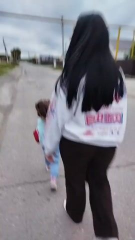 A young girl with long brown hair, wearing a light blue sweatshirt and blue jeans, walks away from the camera down a paved road in a rural area. She carries a red object in her right hand. In the foreground, a person, partially visible and out of focus, walks in the same direction. The girl is walking on the right side of the road. Houses with fences are visible on both sides of the road, and utility poles line the street. The sky is overcast.
