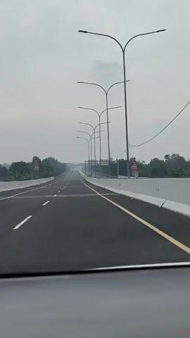 The video shows a view from inside a vehicle traveling on an empty highway under an overcast sky. The highway has multiple lanes with clear white and yellow lane markings. Streetlights line the right side of the road, and a concrete barrier separates the highway from the surroundings. Trees and some structures that resemble minarets are visible on the left side of the highway. The road stretches into the distance, with a few other vehicles visible far ahead. The overall impression is that of a modern, well-maintained highway with minimal traffic.