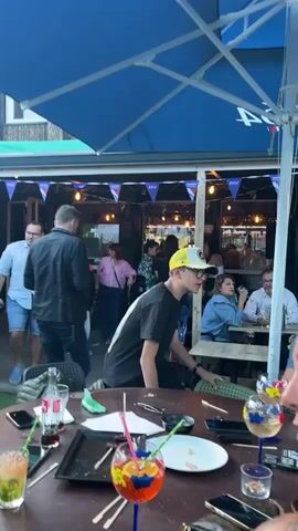 The video appears to be filmed from a table at an outdoor seating area of a restaurant or bar. In the foreground, a woman with long dark hair is partially visible, looking to the right. On the table are two drinks in stemmed glasses with colorful garnishes, a plate, and a smartphone. A young male wearing a black t-shirt and a yellow baseball cap is visible in the mid-ground, looking and gesturing towards the left. Behind him, several people are seated at tables, and others are standing and walking around under a large blue umbrella. A man in a light blue shirt and denim shorts is walking towards the left in the background. The environment appears to be daytime with artificial lights visible in the background.