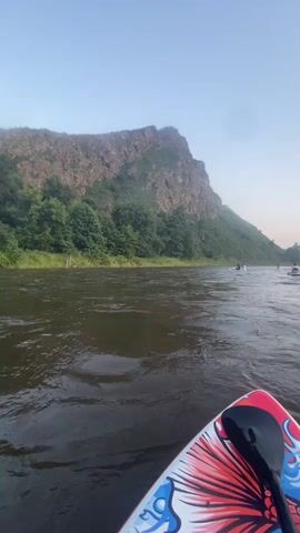 The video shows a first-person perspective from a paddleboard on a river. The paddleboard has a colorful design featuring a fish. In the foreground, a paddle is visible. The river is wide and has a moderate current. On the far bank of the river, there is a steep, rocky mountain covered in trees. The sky is clear and appears to be either early morning or late evening. In the distance, on the river, several other people on paddleboards can be seen. The overall scene is one of outdoor recreation in a natural landscape.