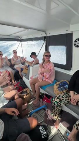 Here is a description of the video:

The video shows the interior of a boat with several people on board. There are five women seated on one side, wearing summer clothing and sunglasses. At least two men are seated across from them, with others partially visible. Everyone is clapping their hands in unison, seemingly enjoying themselves. Outside the boat, the ocean water is visible, with some splashing against the side. The boat is moving at a moderate speed, as indicated by the wake trailing behind. Personal belongings such as bags and beach towels are scattered around the seating area. The interior of the boat is well-lit, with white and gray seating and black accents.