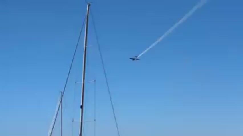 The video shows a sailboat on water under a clear blue sky. An airplane is visible in the sky leaving a contrail. The sailboat has a tall mast and several people on deck. In the distance, a pier and buildings are visible, as well as other boats.
