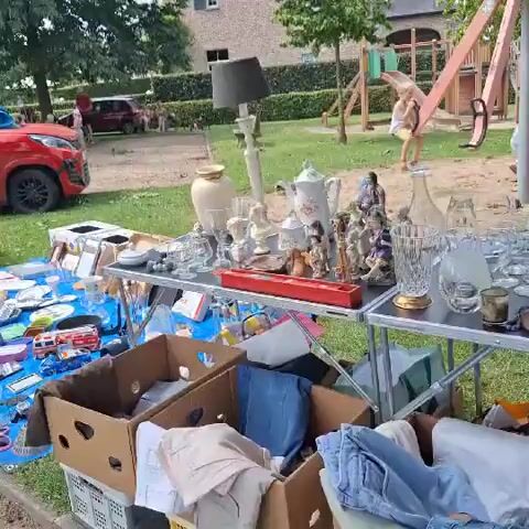 This is a video of a yard sale in progress. In the foreground, items are laid out on tables and on a blue tarp. These items include glassware, vases, figurines, picture frames, children's toys, and miscellaneous household items. In the background, a playground can be seen with several children playing. Two adults are visible in the background, one standing and another sitting on the playground equipment. The setting appears to be a residential backyard with a lawn and trees. The video pans across the yard sale items, showing the variety of goods for sale.