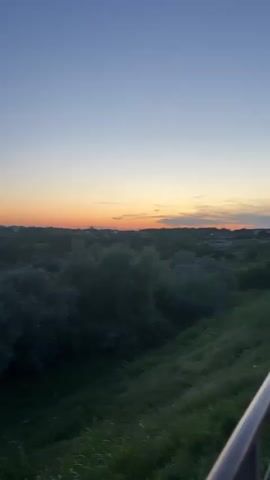 The video shows a panoramic view of a landscape transitioning from a wooded area to the sea at dusk. The sky is a gradient of deep blue at the top fading to orange and pink near the horizon. In the foreground, there is green grass and foliage. The middle ground is dominated by dense trees, some with lighter foliage. Further back, rolling hills covered in vegetation lead to the coastline. The sea is visible on the right side of the frame, appearing calm and reflecting the soft colors of the sky. There are no people or discernible man-made structures in the visible portions of the landscape. The camera appears to be moving horizontally, panning across the scene.