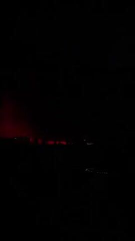 The video shows a dark outdoor scene. Initially, there are vague silhouettes of people standing in front of a rocky cliff, illuminated by a white light. Then, the cliff is lit up with purple light, followed by a shot of a car moving away with its red taillights visible. The video ends with a bonfire in the foreground and indistinct dark shapes in the background.