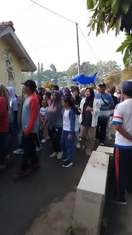 A group of people are walking in a line on an outdoor paved area. The people are of various ages and genders, many wearing casual clothing. Some individuals are wearing headscarves. In the background, there are trees, a building, and a blue shark-shaped balloon. The weather appears to be sunny. The line of people is moving from left to right in the frame. One person in the foreground on the right is wearing a white shirt with red and blue accents.