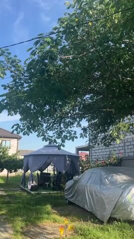 The video shows a backyard scene on a sunny day. The camera pans upwards, focusing on the leaves of a large tree that casts dappled shade over the area. Below the tree, a gray gazebo with outdoor furniture is visible, suggesting a space for relaxation and gatherings. To the right of the gazebo, a car is covered with a silver tarp. In the background, a house constructed of white brick is partially visible, with windows reflecting the blue sky. Pink flowers are also blooming near the house. The sky is clear and blue with some wispy clouds, indicating pleasant weather. The presence of the gazebo, covered car, and house suggests a residential setting, likely a private home. The time of day appears to be daytime, given the bright sunlight and shadows. There is no visible text in the video.