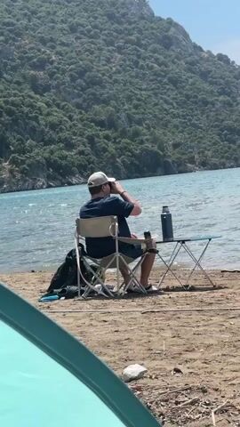 A man is sitting on a folding chair at a small folding table on a sandy beach by the water. He is wearing a dark shirt, shorts, shoes, and a light-colored baseball cap. He has his back to the camera. He takes a drink from a dark-colored thermos on the table. Behind him is a turquoise body of water and a large, green, forested hill. In the foreground, partially obscuring the left side of the frame, is a turquoise tent.